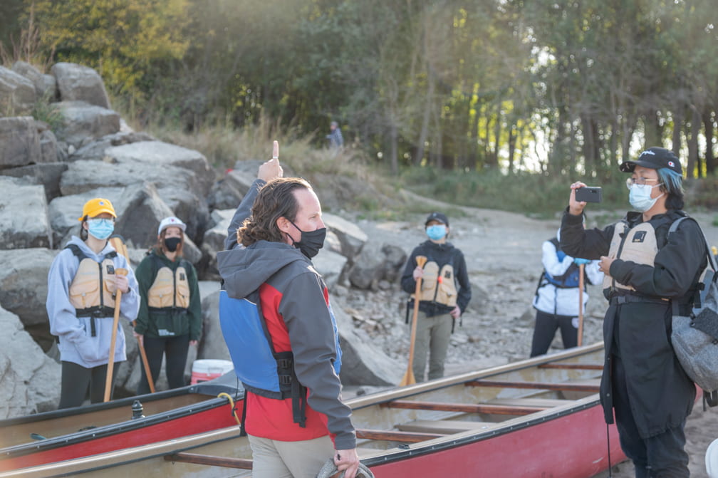 A person in a facemask, red jacket, and lifevest stands in front of two red canoes on a rocky beach, pointing upward. Five people in life jackets look towards them, one films them with a phone.